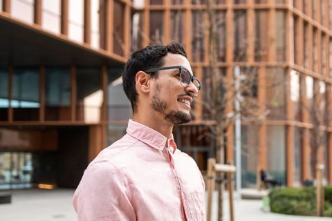 A stylish Cuban man wearing glasses and a light pink shirt smiles confidently. The modern urban