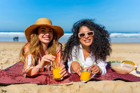 Two smiling multiracial women wearing summer hats and sunglasses relaxing on a beach blanket,