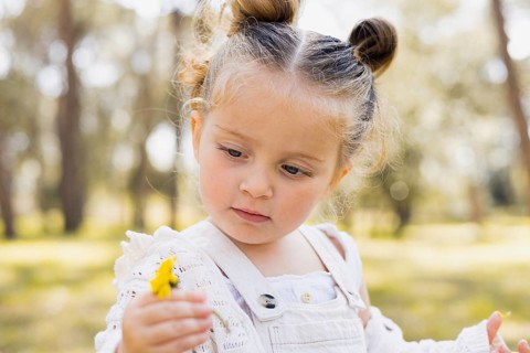 A young girl outdoors in a sunny park, holding and examining a yellow flower with a curious