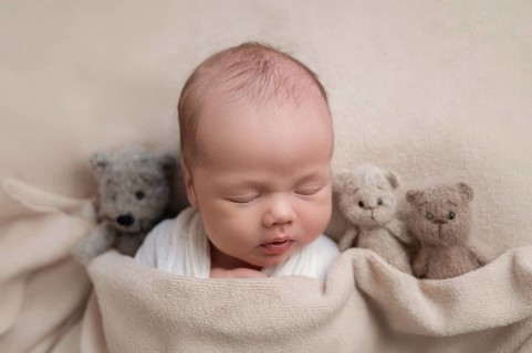 A serene newborn baby sleeps surrounded by three cuddly teddy bears on a soft beige blanket,