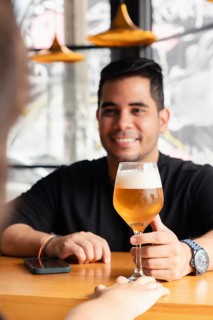 Selective focus on a glass of lager beer with a smiling Latino man in the background receiving the
