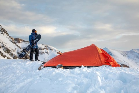 A skier stands next to an orange tent on a snowy mountain, holding a snowboard. The serene winter