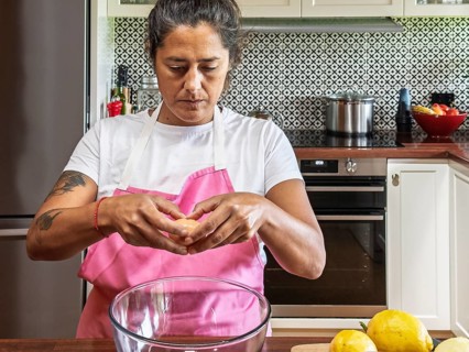 Woman in a pink apron cracks an egg into a glass bowl in a modern kitchen. Lemons and other kitchen