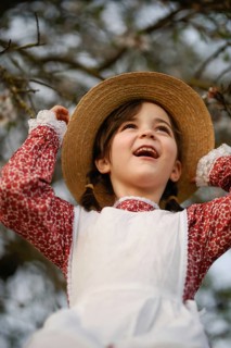 A smiling young girl wearing a straw hat and vintage dress looks up joyfully, surrounded by trees,