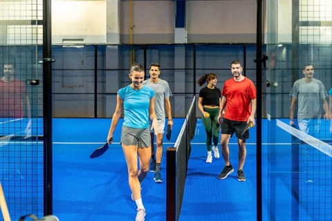 Four friends enter a bright blue pickleball court carrying paddles and balls, smiling and ready to