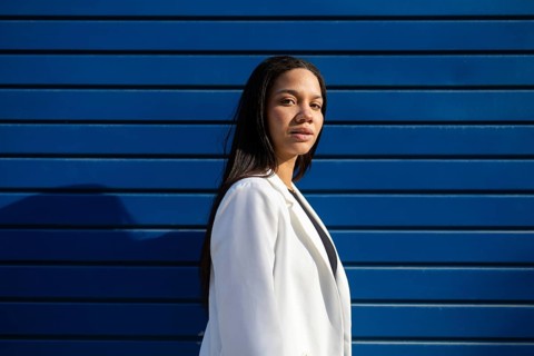 A professional woman in a white blazer stands confidently against a striking blue wall in the