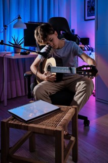 A young boy plays guitar in a cozy, dimly lit room with a microphone. The ambiance includes blue