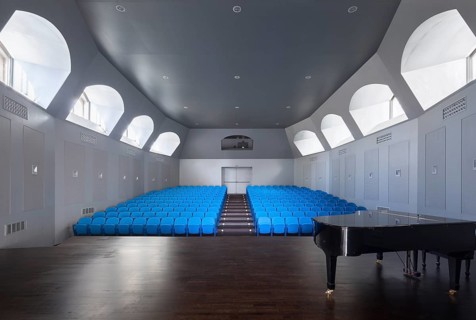 A modern auditorium features rows of blue seats and a sleek black grand piano. The room is well