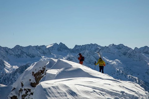 Two freeride skiers traverse a snowy ridge under a clear blue sky, surrounded by stunning mountain