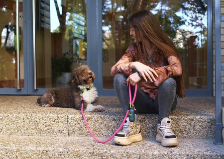 A young woman with long hair sits on steps outside, holding a happy dog on a pink leash. They enjoy