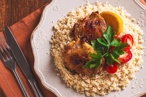 Couscous is served on a plate with pieces of chicken on top. Lemon slices and parsley decorate the