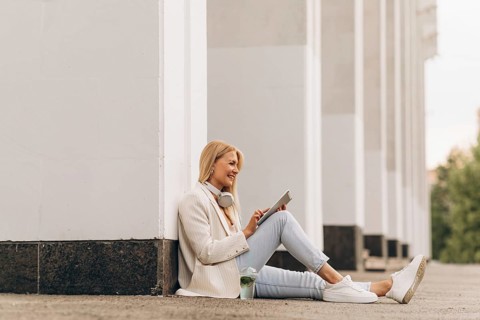 A businesswoman sits against a building, smiling while using a tablet. She wears headphones around