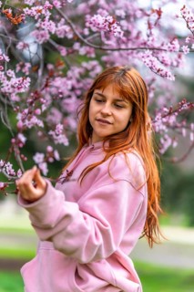 Young woman with long red hair wearing pink hoodie gently touching cherry blossom branch in a park