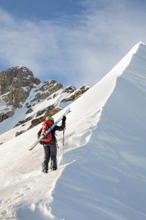 A ski tourer ascends a snow-covered ridge, skis on shoulder, against a backdrop of rugged peaks.