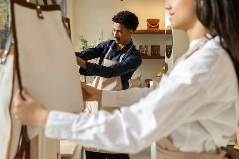 Two young shop assistants wearing aprons are organizing and inspecting bags in a stylish retail