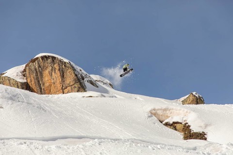 A snowboarder performs a thrilling jump off a snowy cliff, sending powder flying in the backcountry