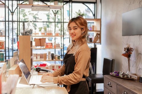 A cosmetics store employee stands by a counter using a laptop, surrounded by skincare products. The