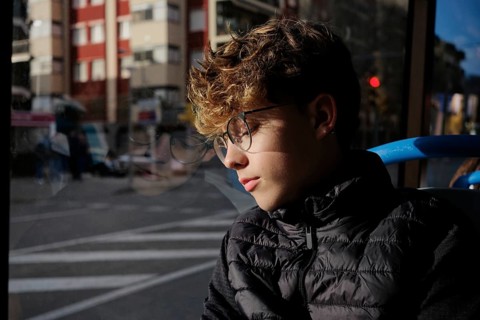A young man wearing glasses and a black jacket sits on a bus, gazing thoughtfully out of the