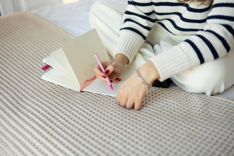 A woman wearing a striped sweater writes in a notebook using a pink pen while sitting on a bed The
