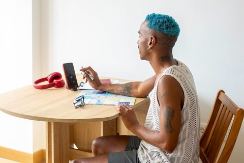 Hispanic LGBTQIA+ man with blue hair planning a holiday trip, sitting at a table with a smartphone,