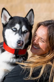 A cheerful woman smiles warmly while holding her husky close. The Siberian husky, wearing a red