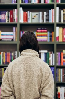 Back view of unrecognizable young girl stands facing a colorful bookcase, engrossed in choosing a