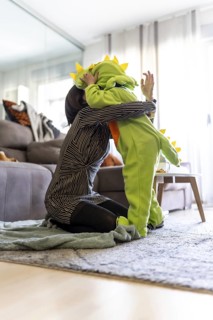 Mother kneeling on carpet and hugging her son wearing a dinosaur costume while playing together in