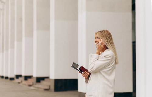 A businesswoman in a white suit stands outside a modern building, engaged in a phone conversation.