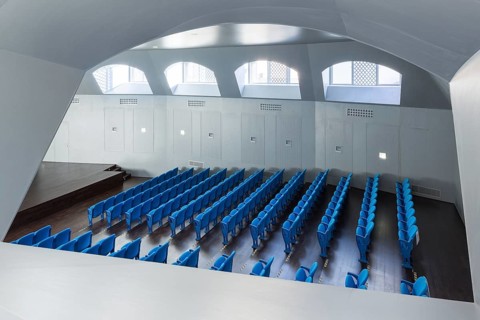 Bright modern auditorium featuring rows of blue seats, wooden flooring, and natural light entering