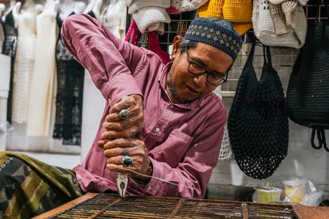 A balinese artisan intensely crafting with pliers and wire, surrounded by vibrant crocheted items