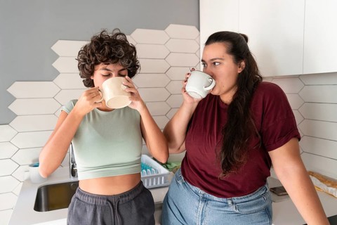 A lesbian couple stands in a cozy kitchen, sipping morning coffee. The modern decor and soft