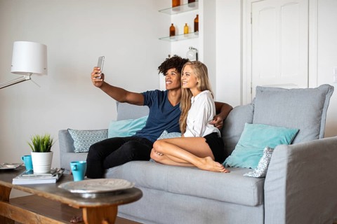 A cheerful couple sitting on a sofa in a modern living room, capturing a selfie with a smartphone.