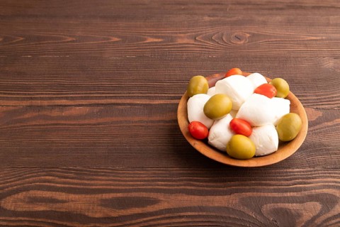 White Mozzarella cheese, with tomatoes and olives in wooden bowl on brown wooden background, side