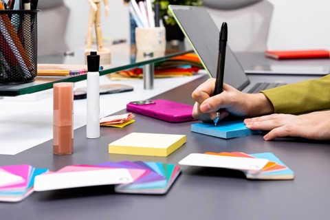 A creative workspace featuring hands writing on a blue notepad surrounded by colorful tools,