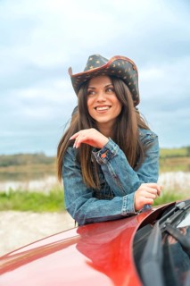Vertical portrait of a smiling woman with cowboy hat leaning on a car in a road