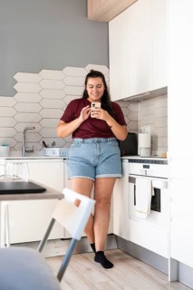 A curvy woman stands in a modern kitchen, smiling while using a smartphone. She leans against the