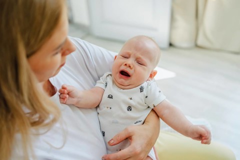 A parent gently comforts a crying baby in a warm, bright living room. The scene captures a tender