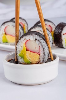 A close up of a sushi roll held by chopsticks, dipping into a soy sauce bowl in white background.