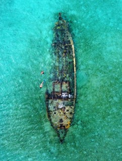 Aerial view of two unrecognizable people, swim around a moss-covered sunken ship in the clear,