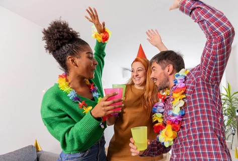 A lively gathering of friends celebrating a birthday with colorful leis, party hats, and drinks.