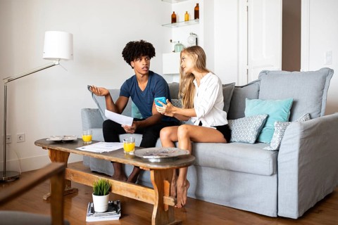 A couple is seated on a cozy gray couch, engaging in a lively discussion while holding papers and a