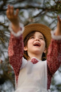 A cheerful girl in vintage clothing and a straw hat, reaching upward with joy against a blurred
