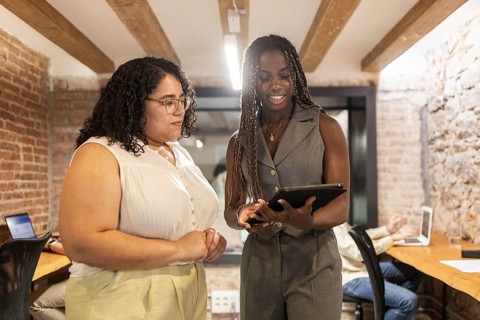 Two women are engaged in discussion, sharing ideas over a tablet in a modern coworking space. A