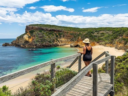 A woman in a hat and swimsuit stands on a wooden walkway, admiring a serene coastal scene with