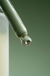 Macro shot of a cosmetic serum dropper with a single golden drop hanging at the tip against a muted