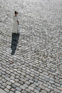 A young girl stands alone in a white First Communion dress, casting a long shadow on a cobblestone
