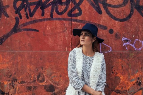 A woman wearing a fashionable black hat and casual outfit leans against a vibrant graffiti-covered