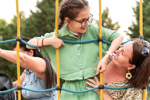 A family enjoys a sunny day at a playground. The focus is on a smiling girl with a tracheostomy,