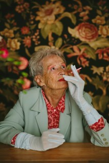 An elderly woman in a vintage setting, wearing elegant attire, looks thoughtfully with a pink