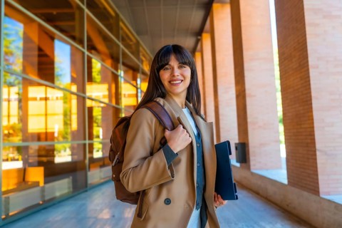 Young woman carrying a backpack and tablet, confidently smiling while walking on a modern